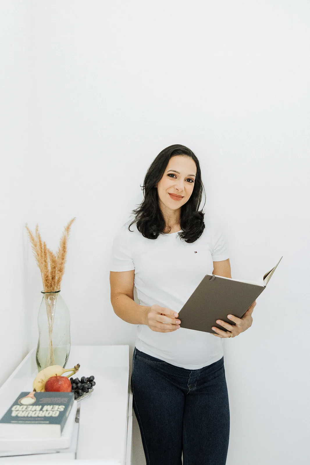 Ana Paula Costa, grávida, de camiseta branca e jeans, segura um caderno aberto e sorri em um consultório claro com frutas sobre a bancada.