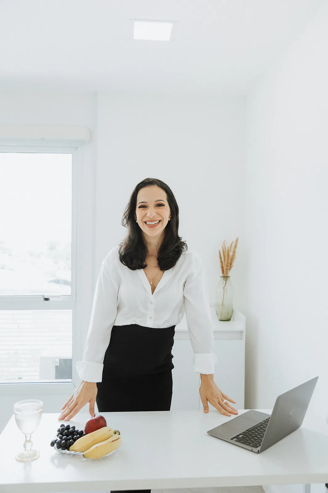 Ana de camisa branca e saia preta apoia as mãos na bancada branca do consultório, com notebook, frutas e um copo de água.