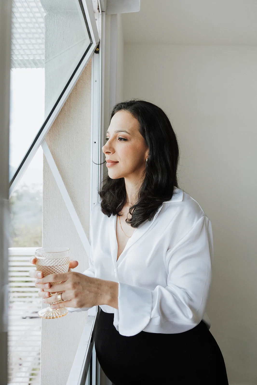 Ana de camisa branca, de perfil junto à janela, segura um copo de água e olha para fora.