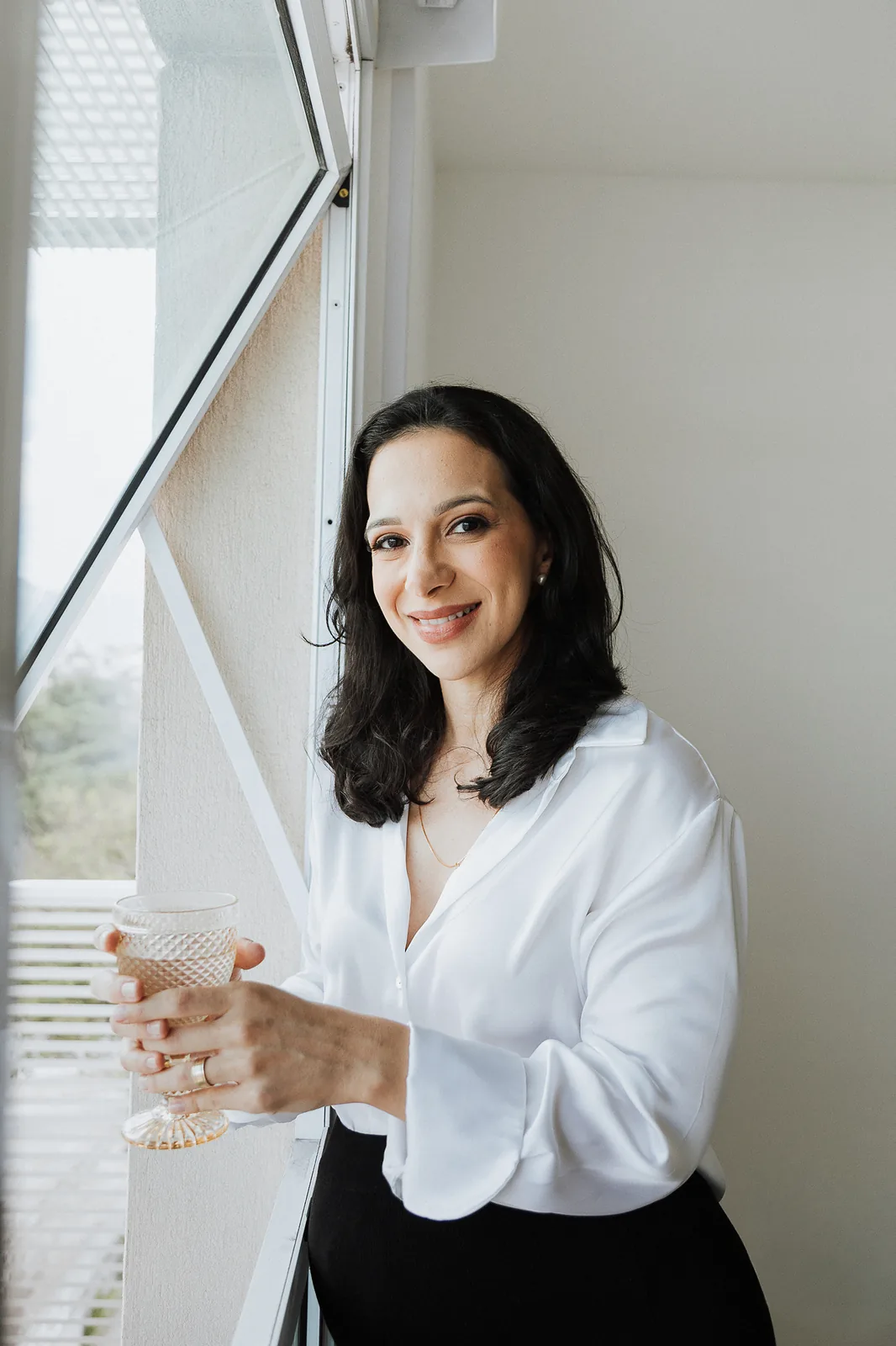 Ana Paula Costa, de camisa branca, junto à janela, segura um copo de água e sorri para a câmera.