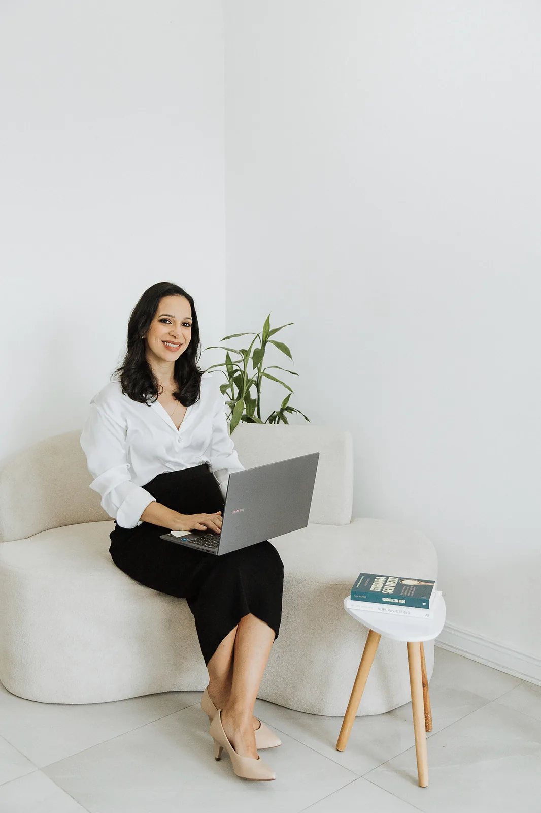 Ana sentada em um sofá claro com o notebook no colo, ao lado de uma planta e de uma mesinha com livro.