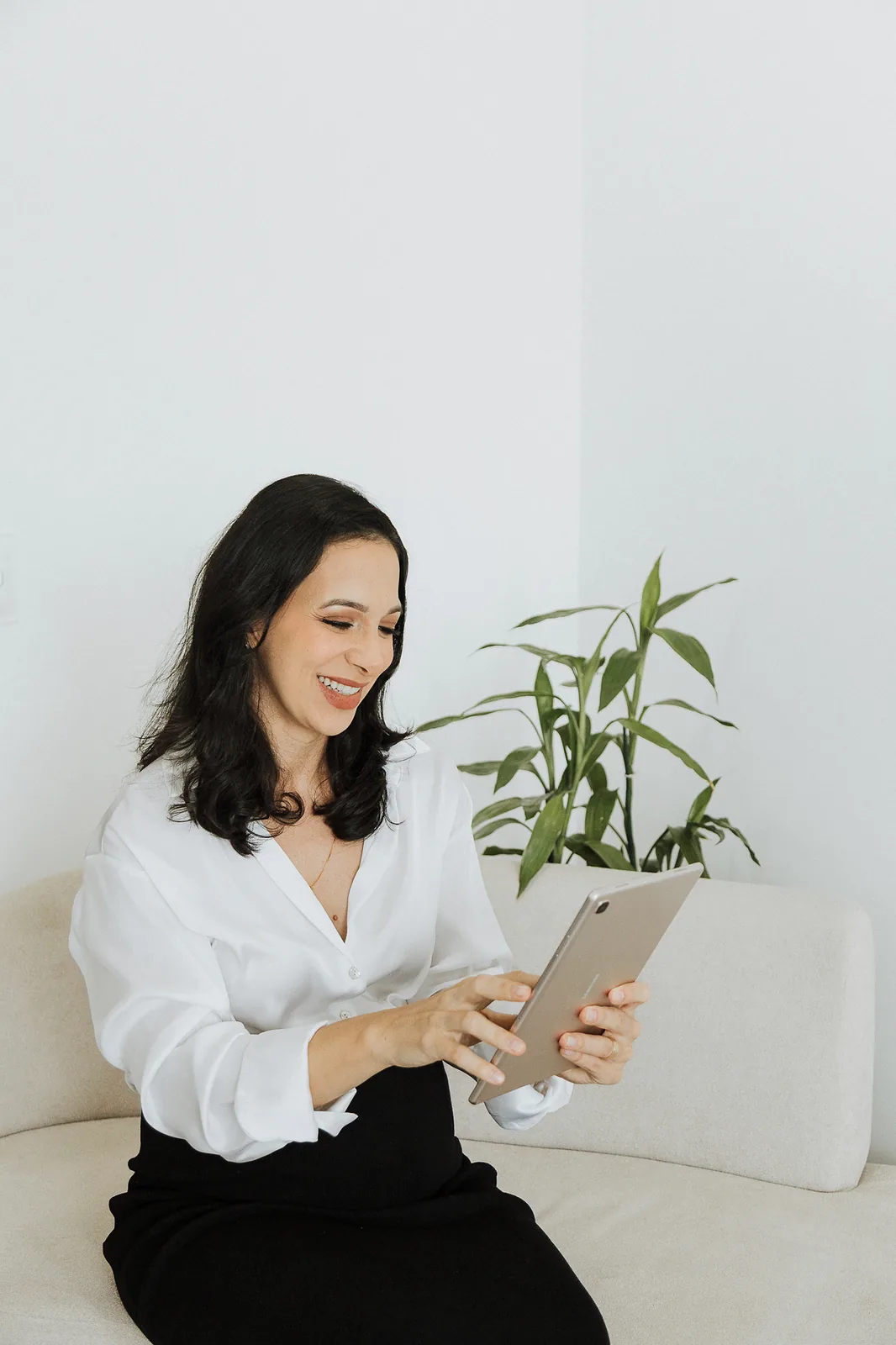 Ana sentada no sofá, de camisa branca, olha para um tablet nas mãos e sorri.