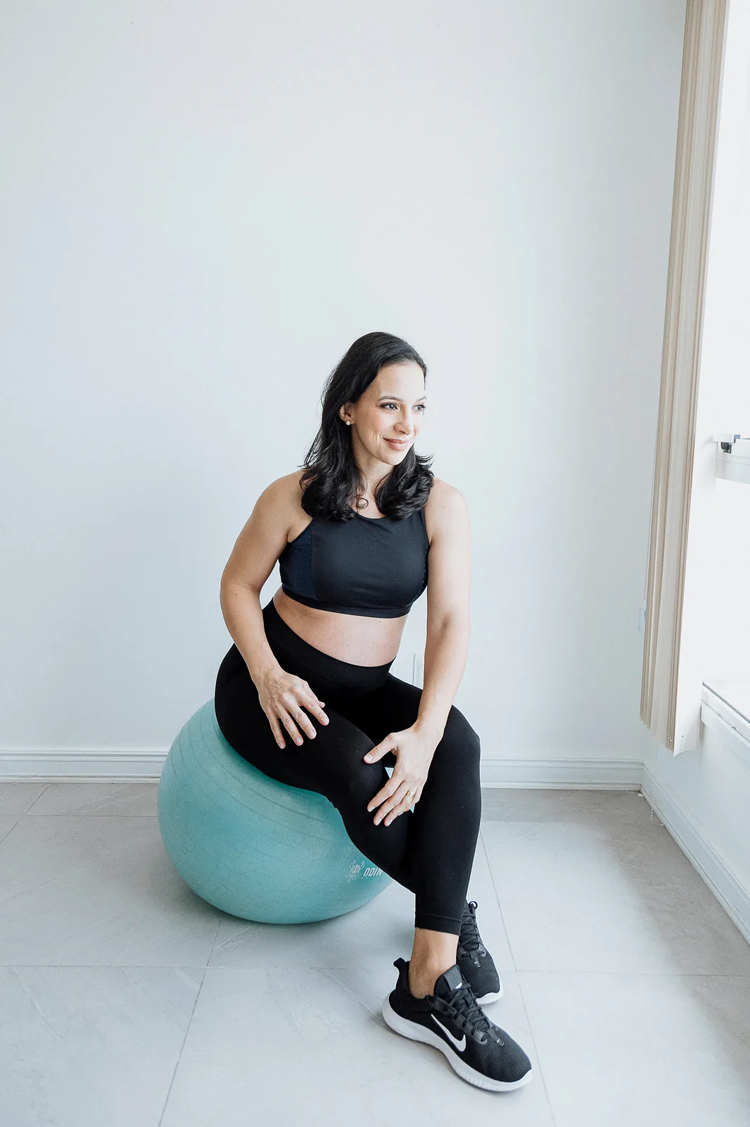 Ana, grávida, de top e legging pretos, sentada em uma bola de pilates azul-esverdeada, sorri com as mãos nos joelhos.