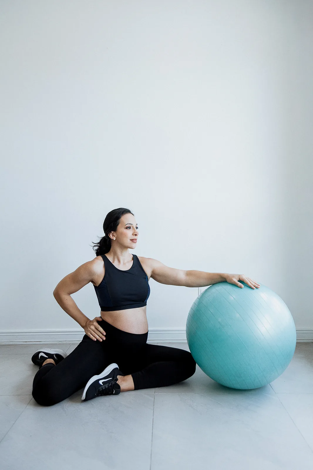Ana, grávida, de top e legging pretos, sentada no chão em alongamento com o braço apoiado em uma bola de pilates.
