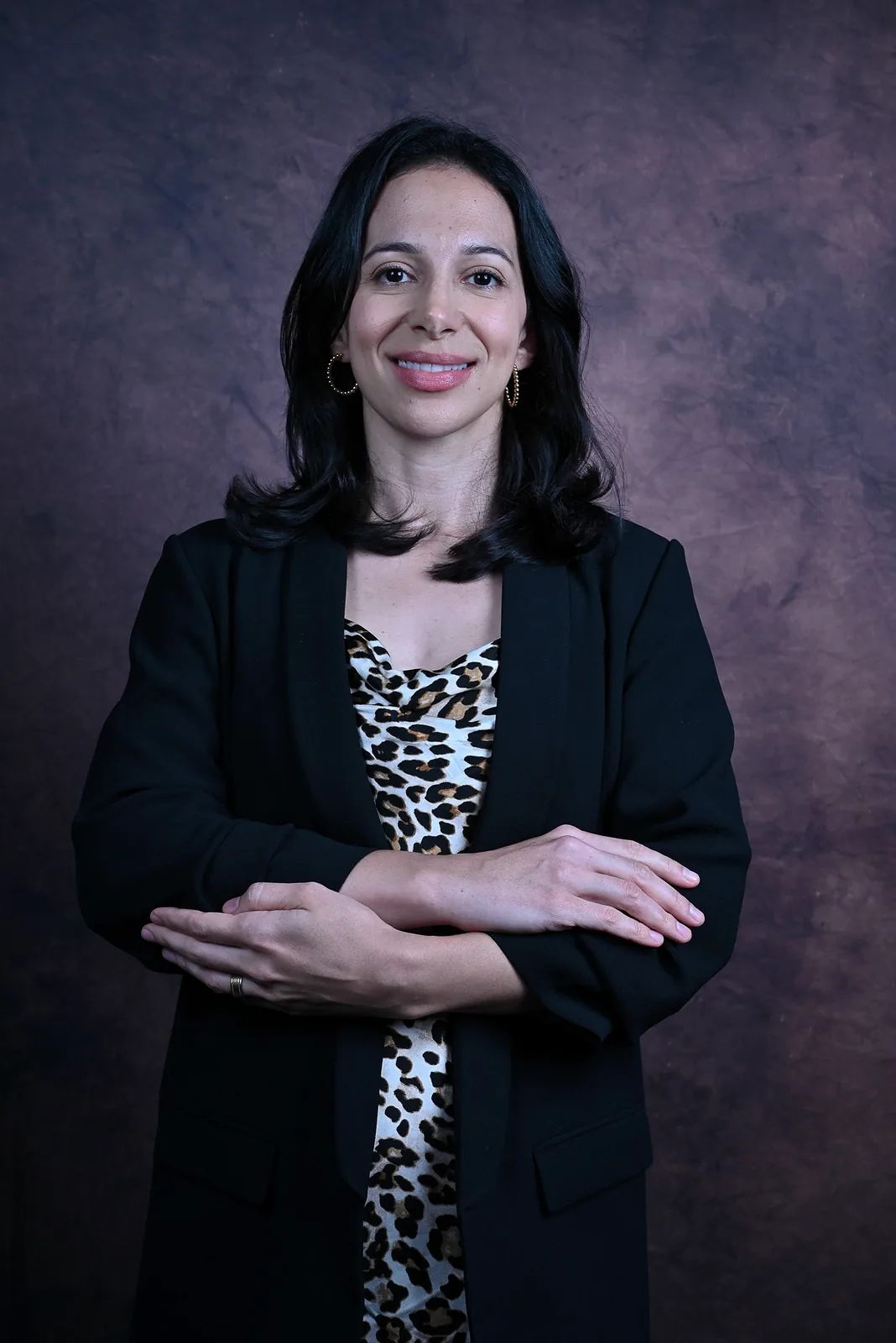 Ana de blazer preto e vestido estampado, de braços cruzados, sorri diante de um fundo escuro texturizado.