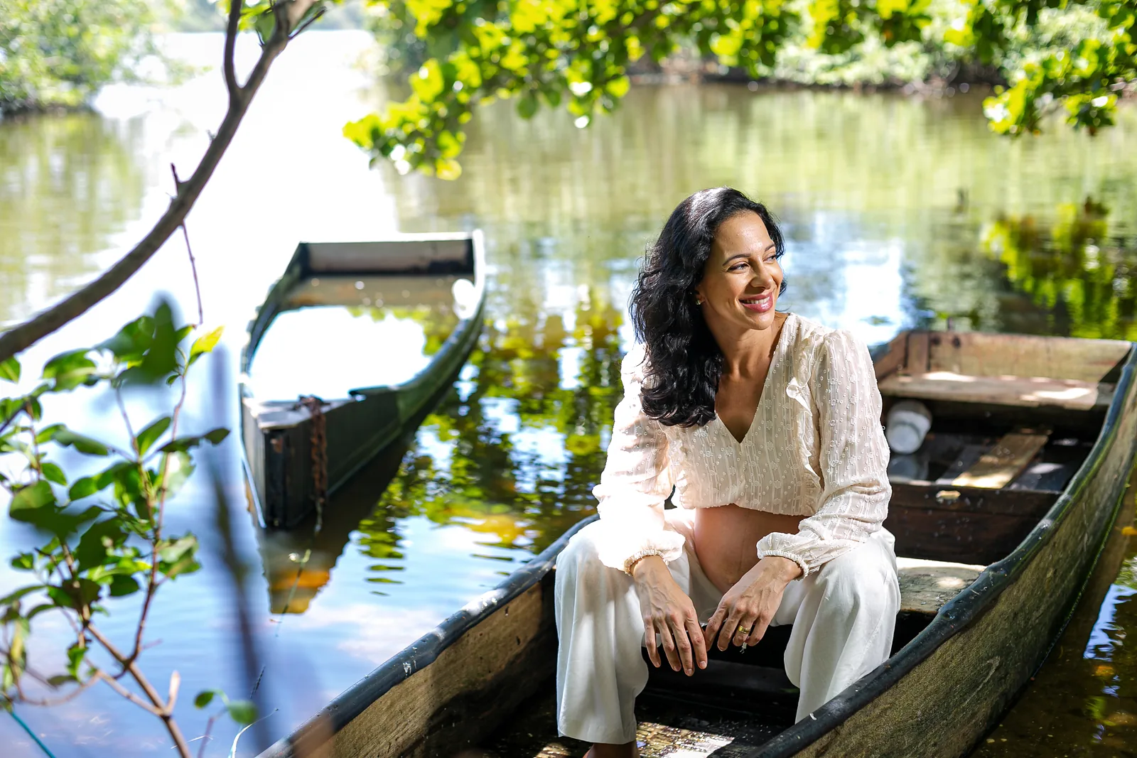 Ana, grávida, de roupa clara, sentada em um barco de madeira na beira de uma lagoa com árvores, sorrindo.