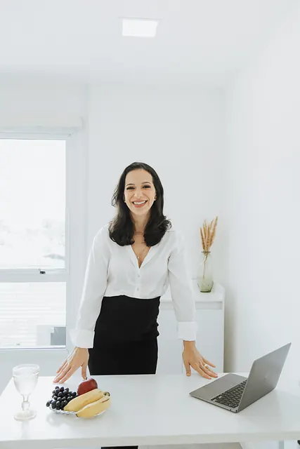 Ana de camisa branca e saia preta apoia as mãos na bancada branca do consultório, com notebook, frutas e um copo de água.
