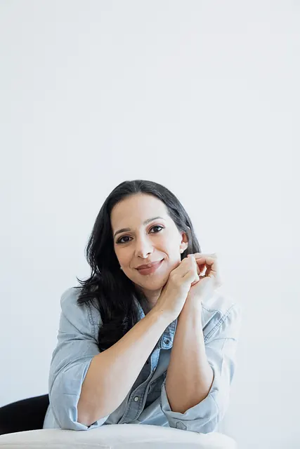 Retrato próximo da Ana de camisa jeans, com as mãos juntas perto do rosto, sorrindo para a câmera.