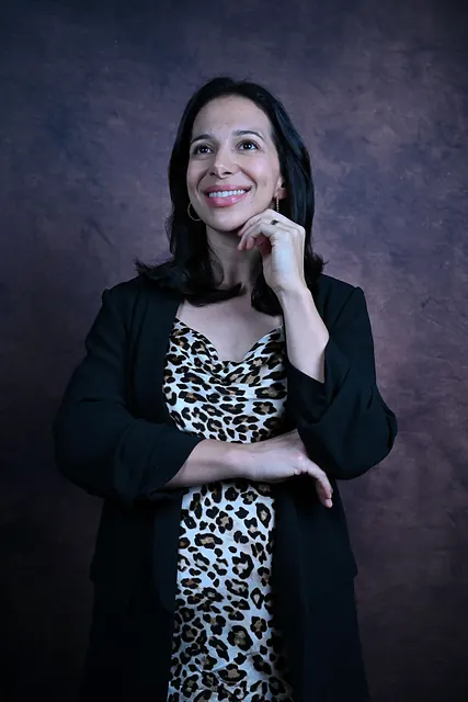 Ana de blazer preto e vestido estampado, com a mão no queixo, sorri diante de um fundo escuro texturizado.
