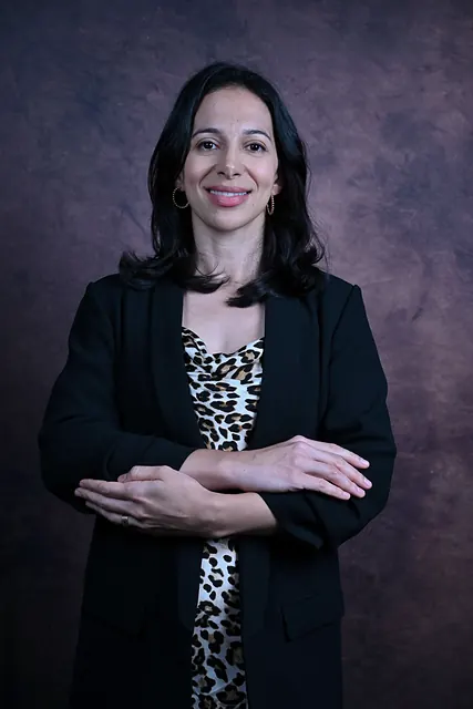 Ana de blazer preto e vestido estampado, de braços cruzados, sorri diante de um fundo escuro texturizado.