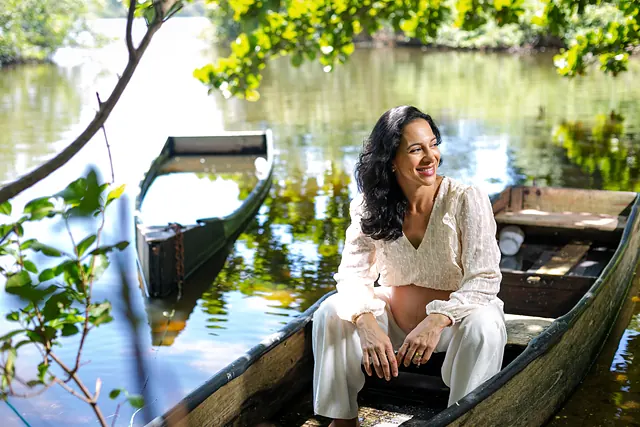 Ana, grávida, de roupa clara, sentada em um barco de madeira na beira de uma lagoa com árvores, sorrindo.