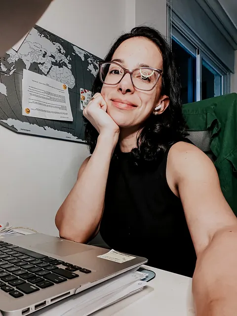 Selfie da Ana de óculos e blusa preta, com o queixo apoiado na mão, diante do notebook e de um mapa-múndi na parede.