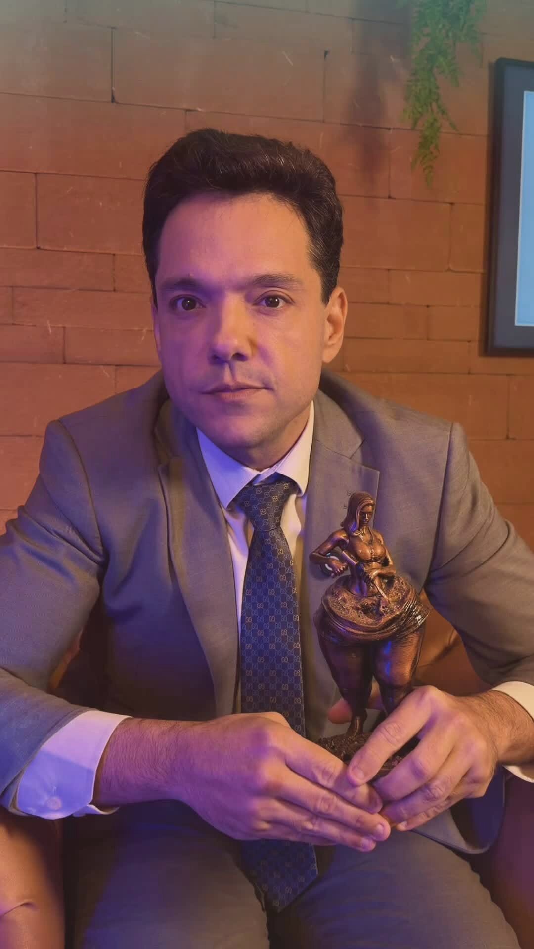 Caico Borelli na poltrona, com luz roxa de um lado e laranja do outro, segurando uma estatueta de bronze