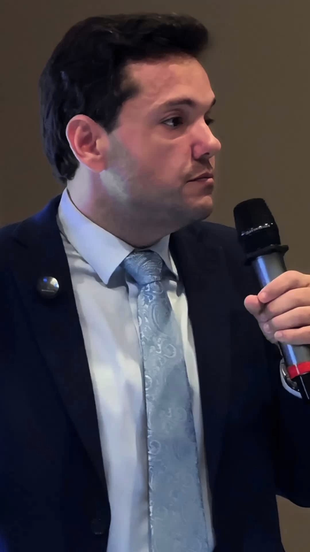 Recorte fechado de Caico Borelli falando ao microfone, de terno azul-escuro e gravata azul-clara, fundo neutro