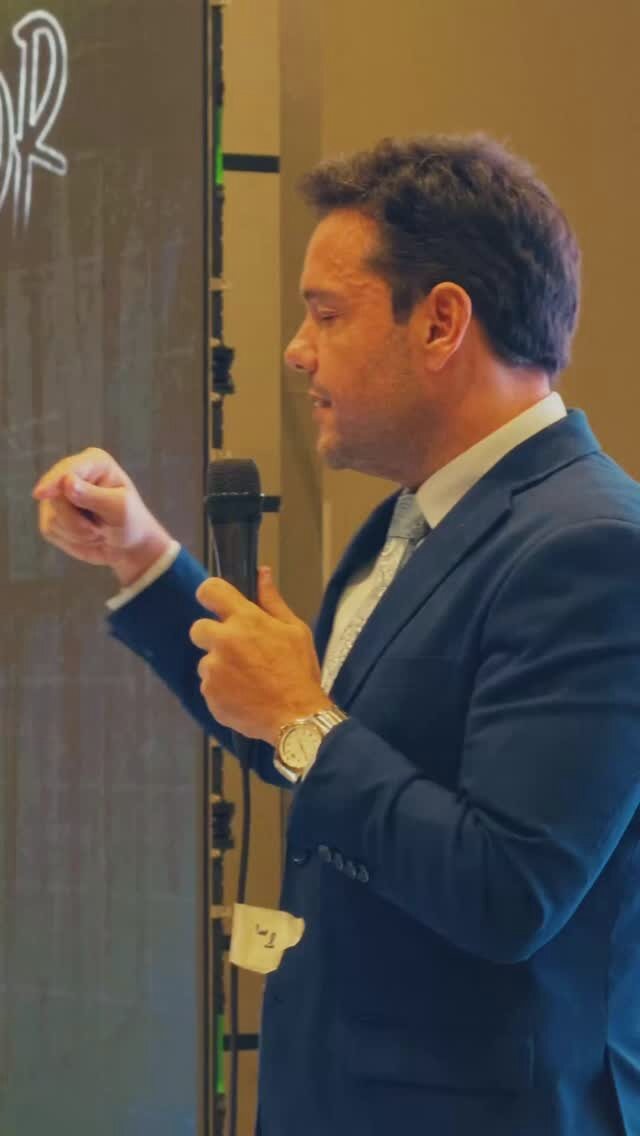 Caico Borelli de perfil numa palestra, com o microfone na mão e luz quente e neutra no rosto