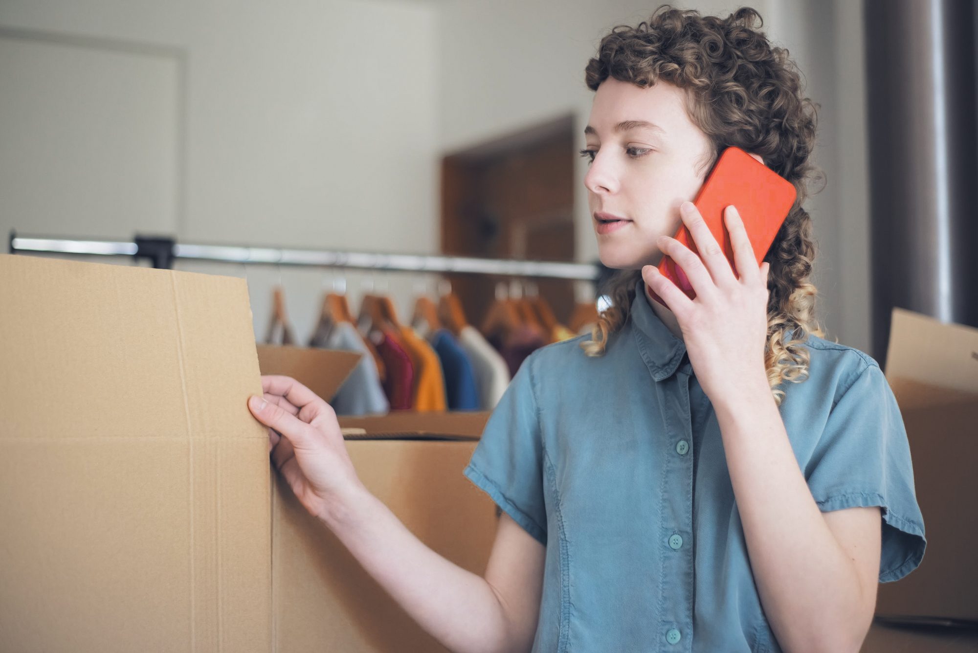 Dona de e-commerce ao celular confere uma caixa de envio no estoque, araras de roupa ao fundo