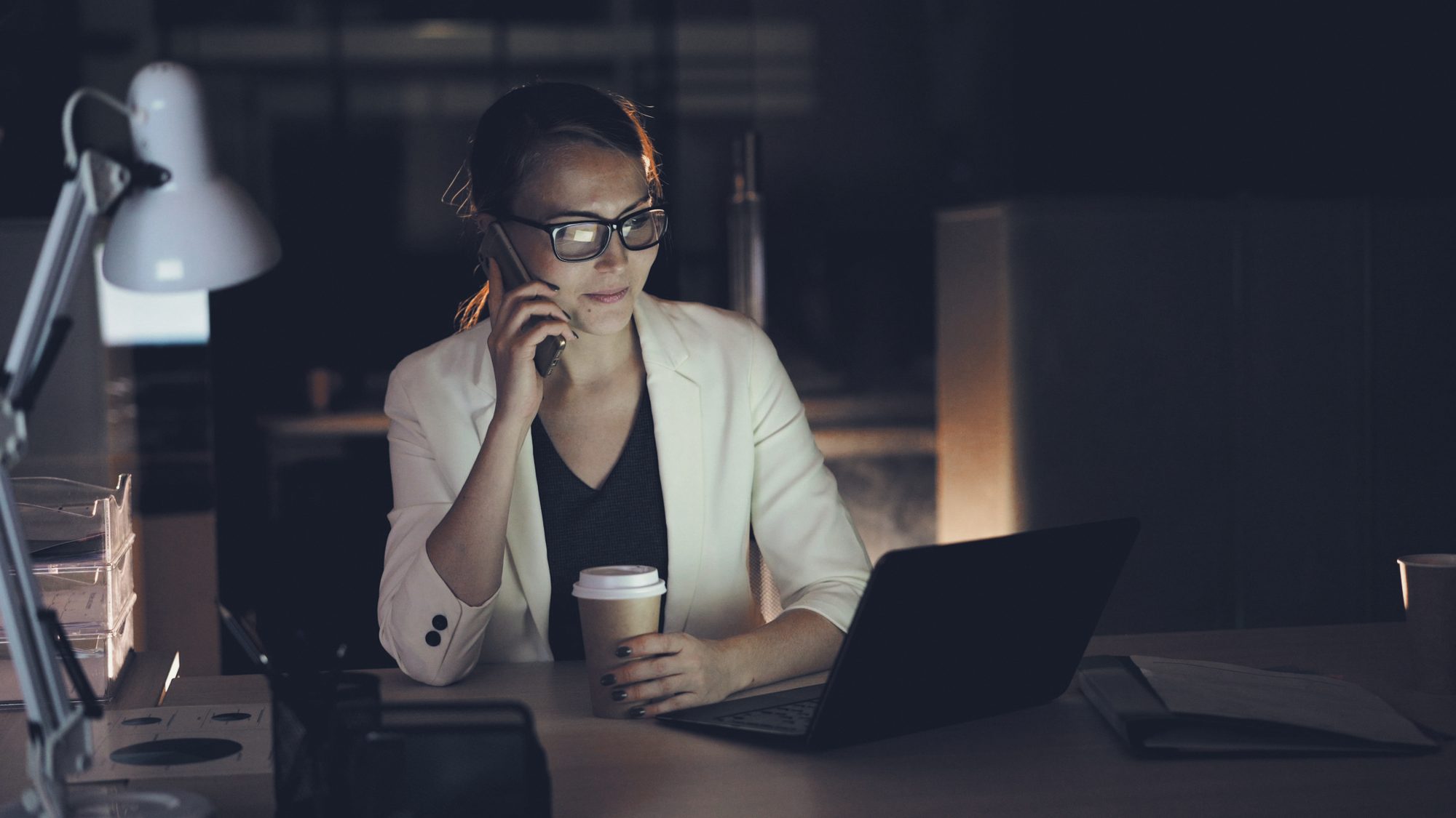 Mulher ao telefone na mesa, notebook aberto, escritório à noite