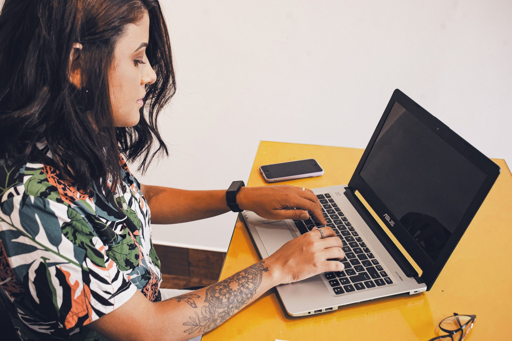 Empreendedora brasileira digitando no notebook numa mesa amarela, olhar na tela