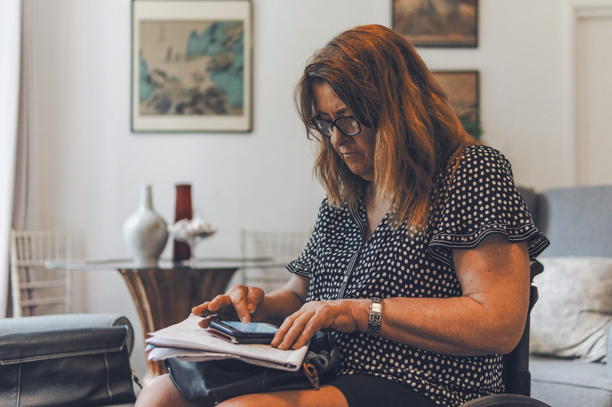 Dona de empresa lendo papéis com o celular na mão, à mesa de casa