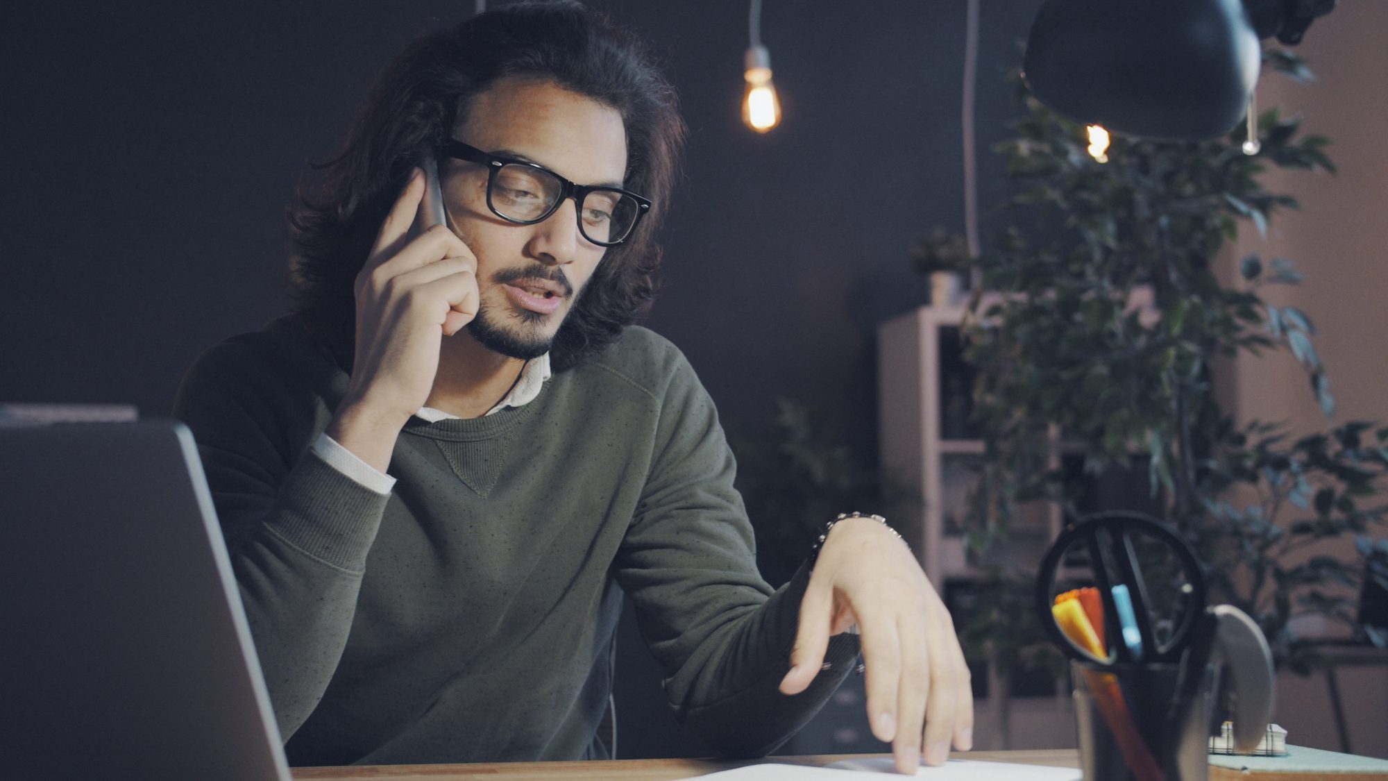 Dono de empresa ao telefone na mesa do escritório à meia-luz, notebook aberto