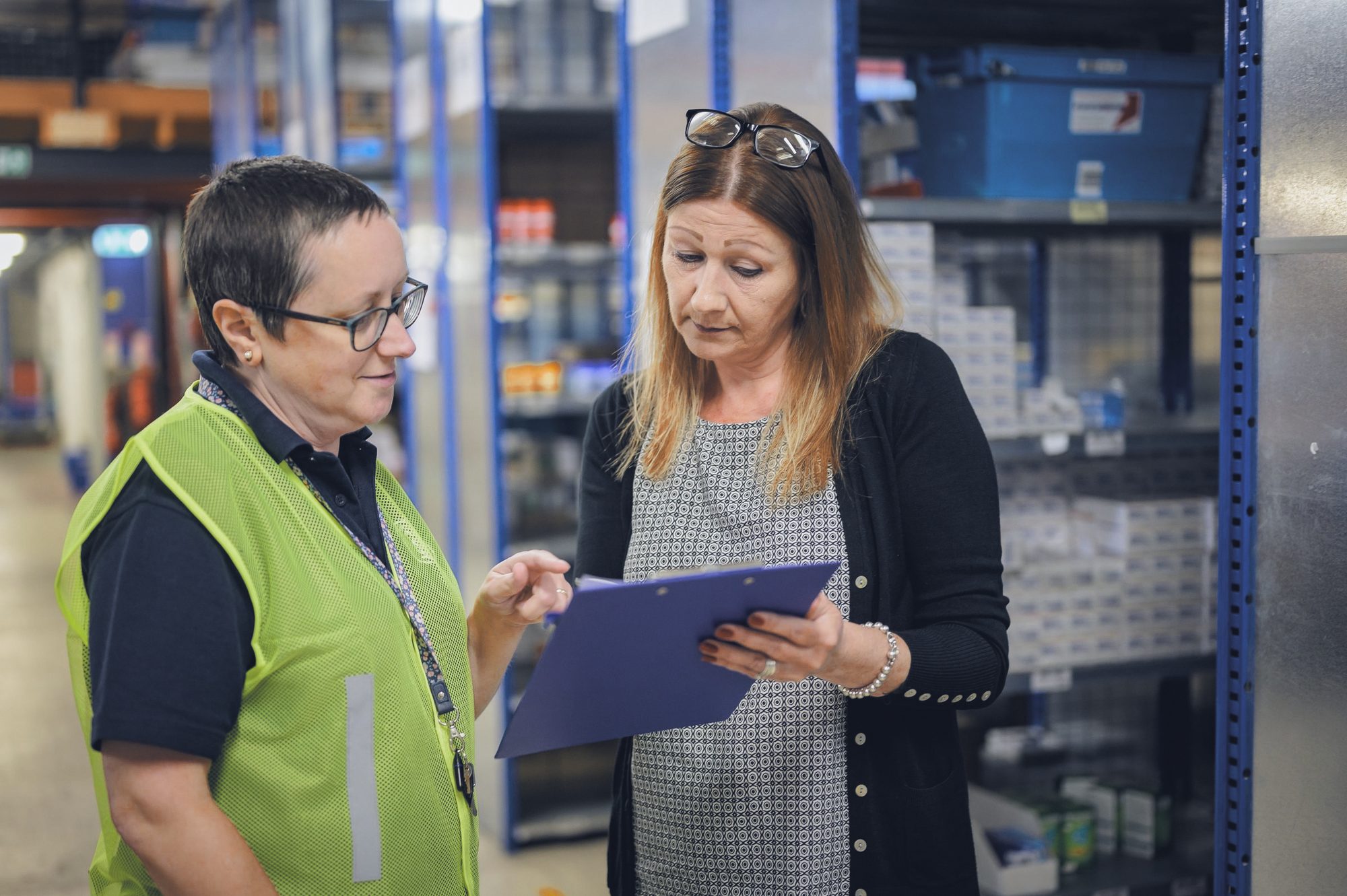 Líder de operação confere a prancheta com uma colaboradora entre as prateleiras do estoque
