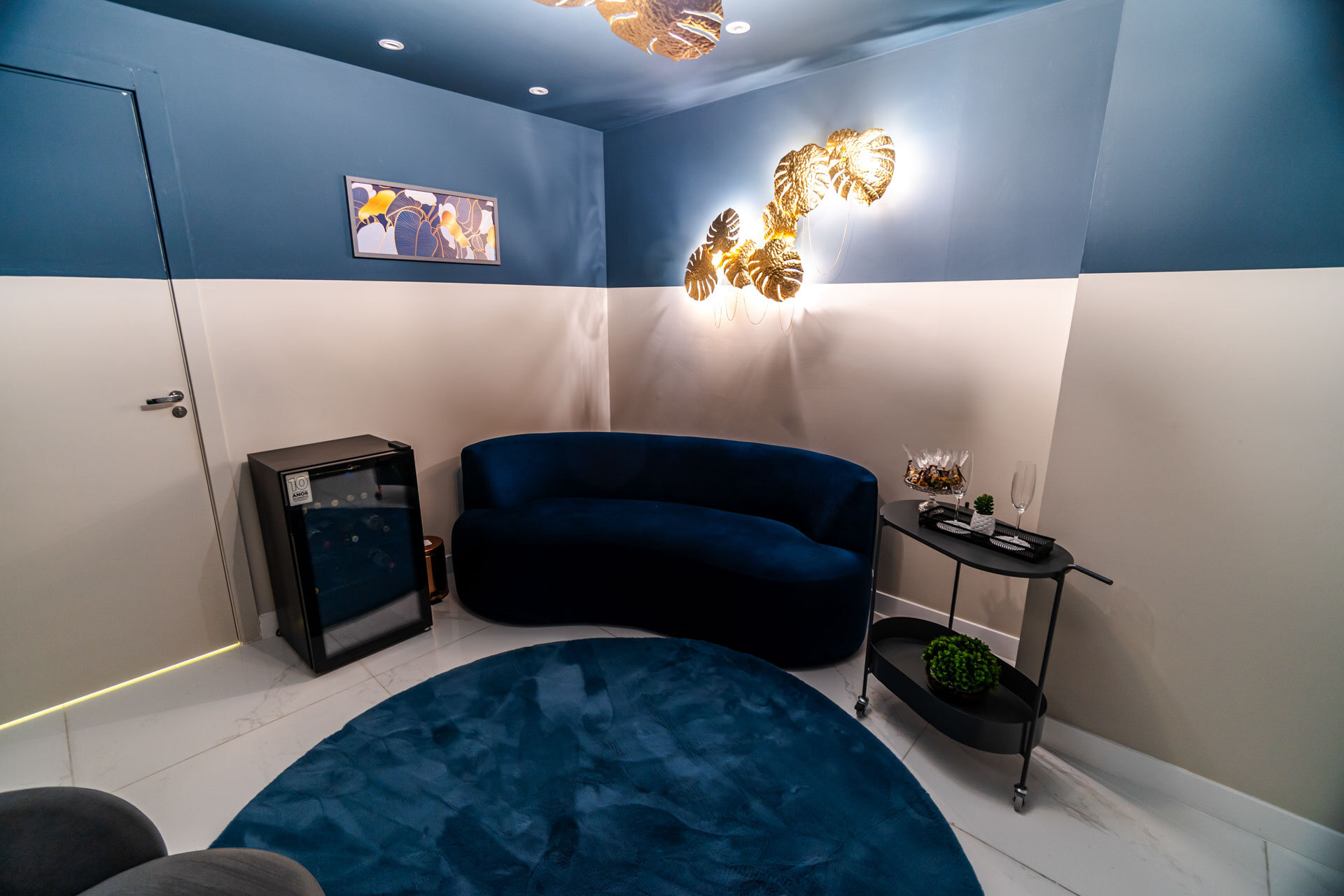 Lounge com parede azul-marinho, sofá de veludo azul e arandelas douradas em forma de folha