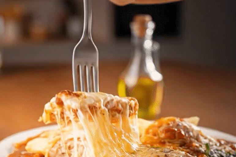 Garfo levantando um pedaço de parmegiana com o queijo puxando, sobre molho de tomate, num sousplat de palha