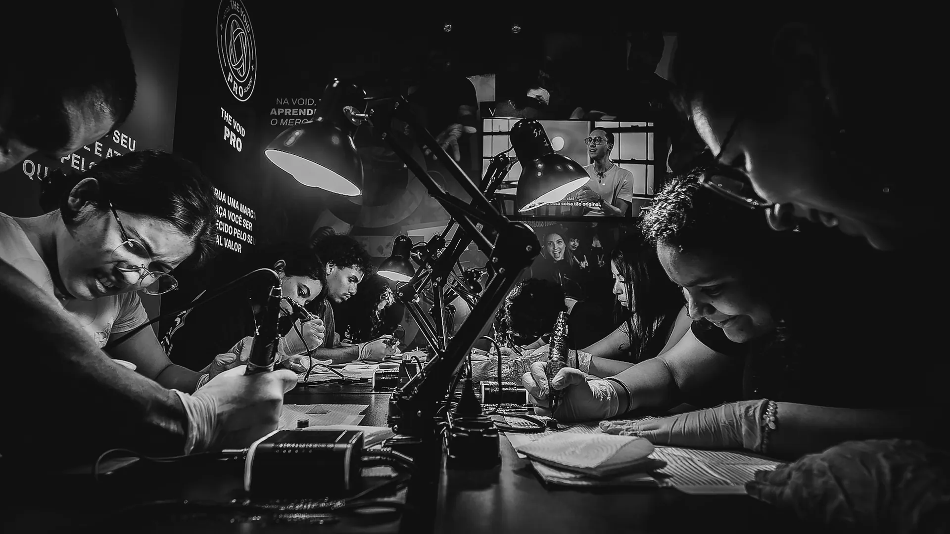 Alunas praticando tatuagem em pele sintética sob luminárias numa mesa coletiva