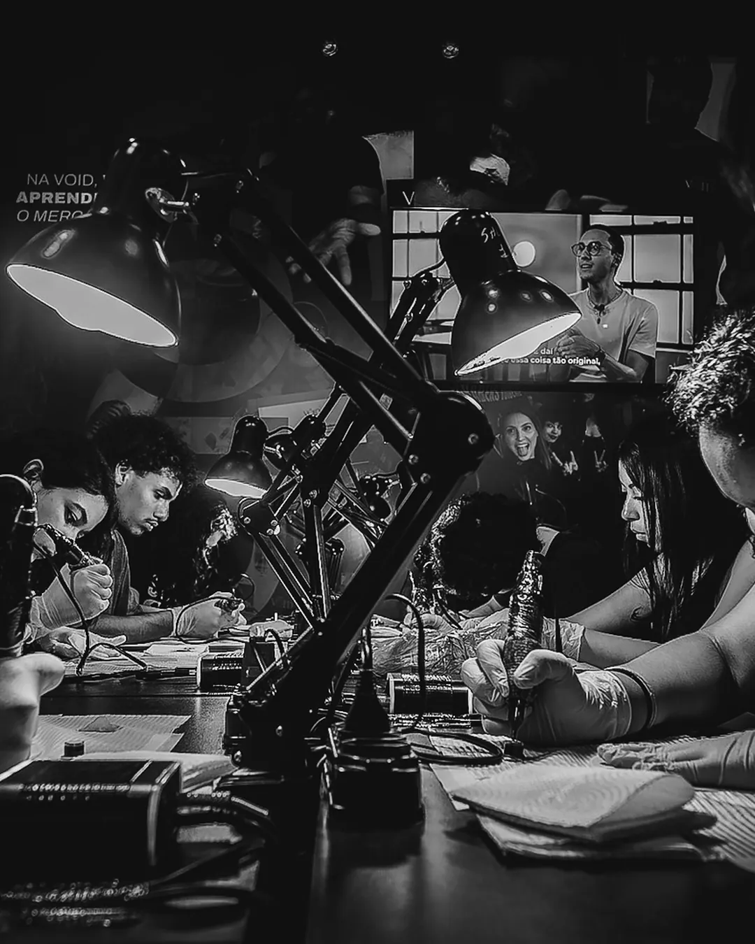 Alunas praticando tatuagem em pele sintética sob luminárias numa mesa coletiva