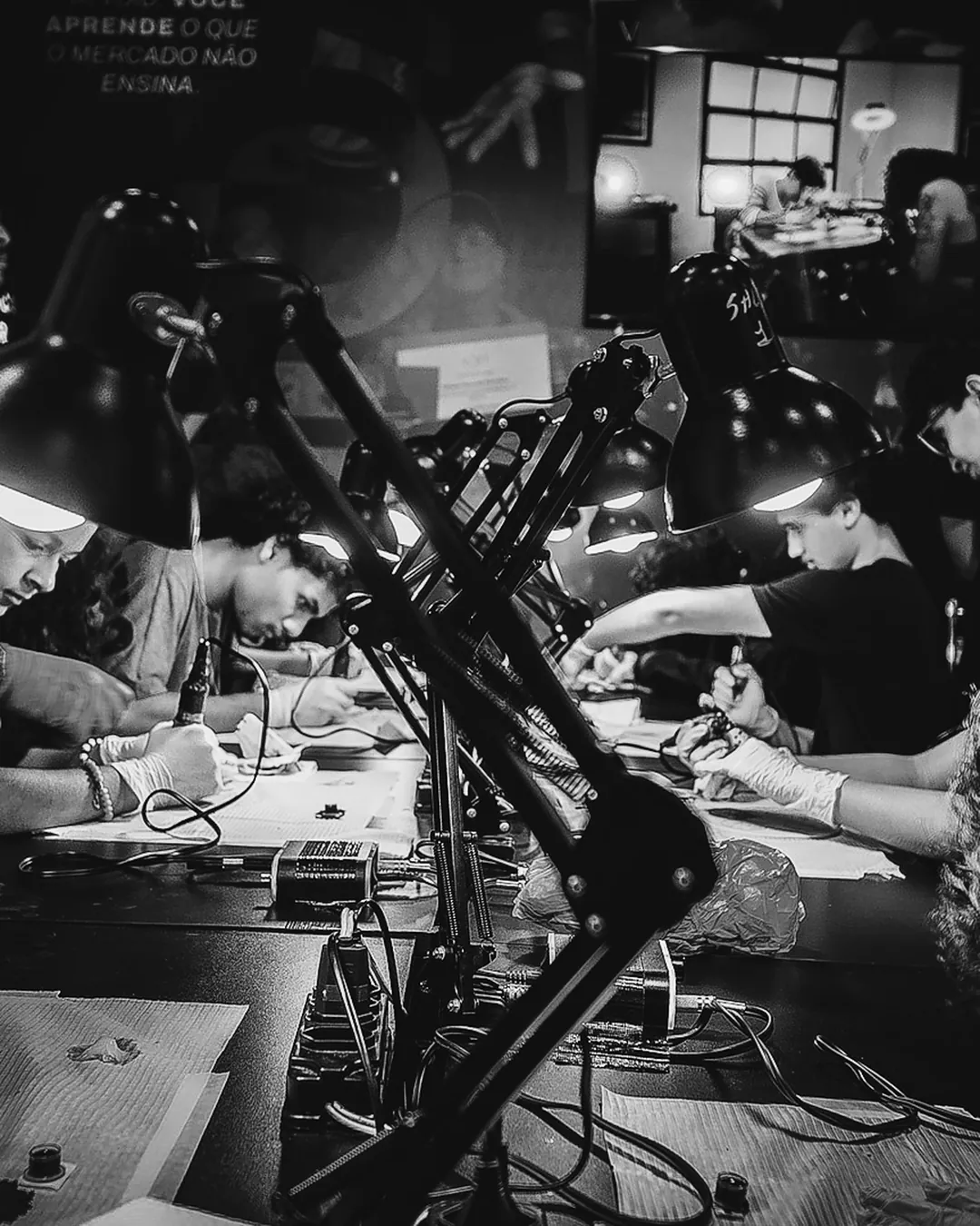 Mesa longa de alunos praticando tatuagem em pele sintética sob luminárias