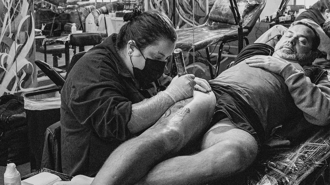 Aluna de máscara tatuando a perna de um cliente deitado na maca, sala com murais da escola