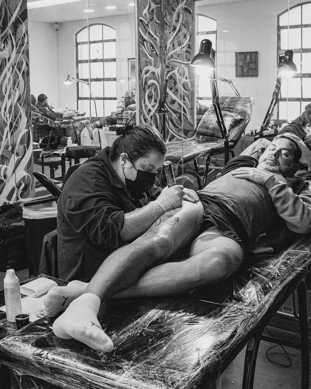 Aluna de máscara tatuando a perna de um cliente deitado na maca, sala com murais da escola