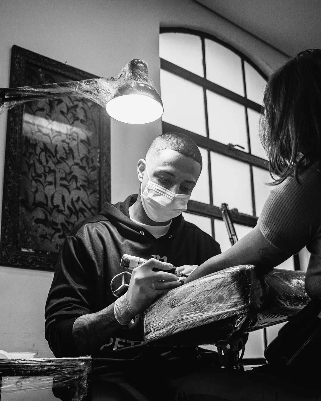 Aluno de máscara tatuando o antebraço de um cliente sob luminária, janelas em arco ao fundo