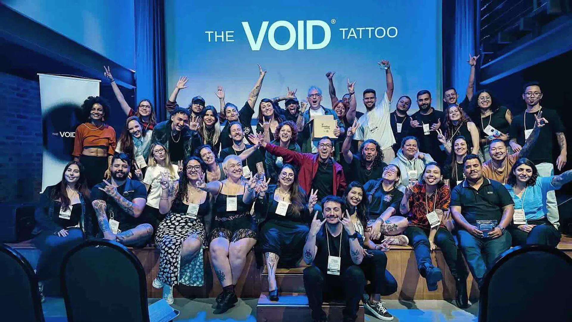Turma grande da The VOID comemorando de braços erguidos sob o telão da escola