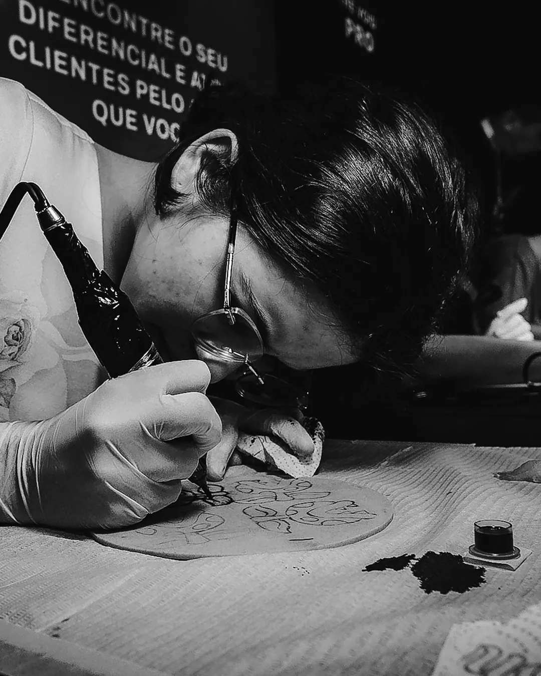Quadro de Reels: aluna de óculos e luvas concentrada tatuando pele sintética sob a luminária
