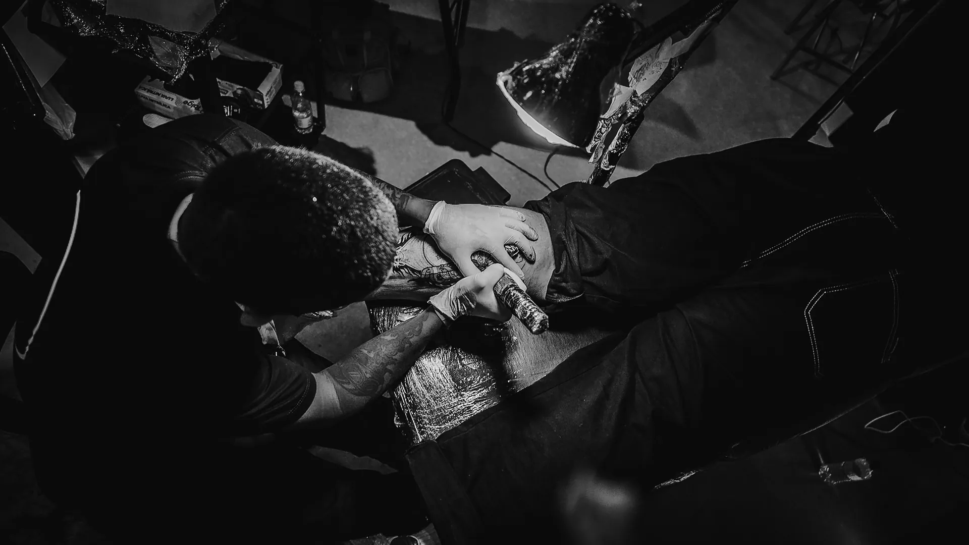 Tatuador debruçado sobre o trabalho sob a luz da luminária, visto de cima
