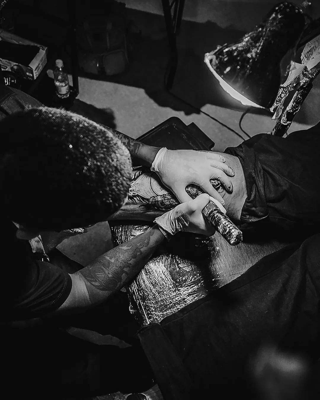 Tatuador debruçado sobre o trabalho sob a luz da luminária, visto de cima