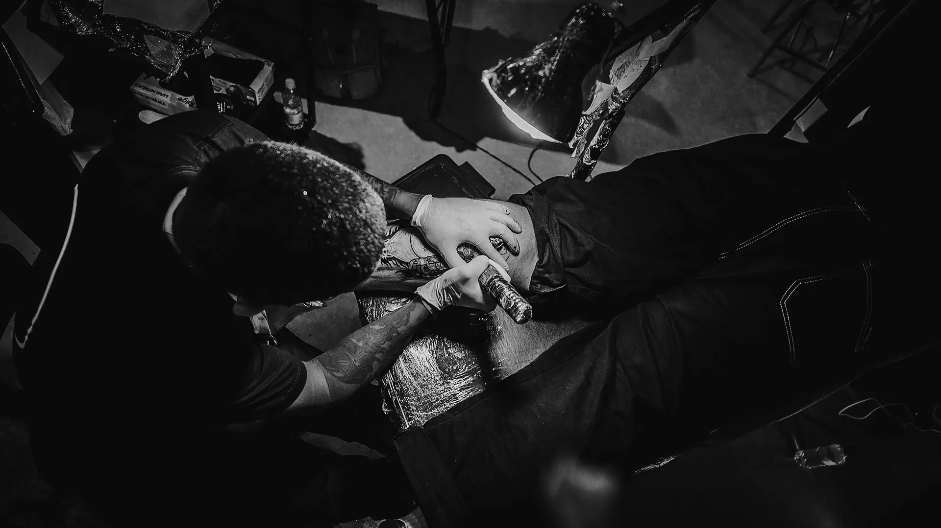 Tatuador debruçado sobre o trabalho sob a luz da luminária, visto de cima