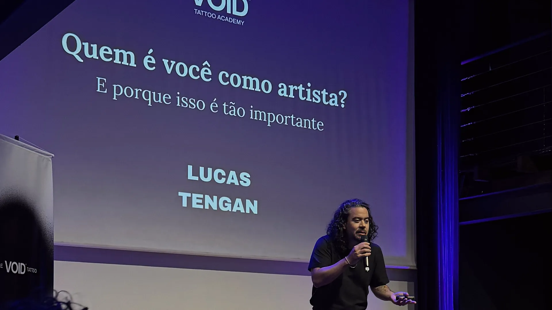Lucas Tengan palestrando no palco, com o telão perguntando quem é você como artista