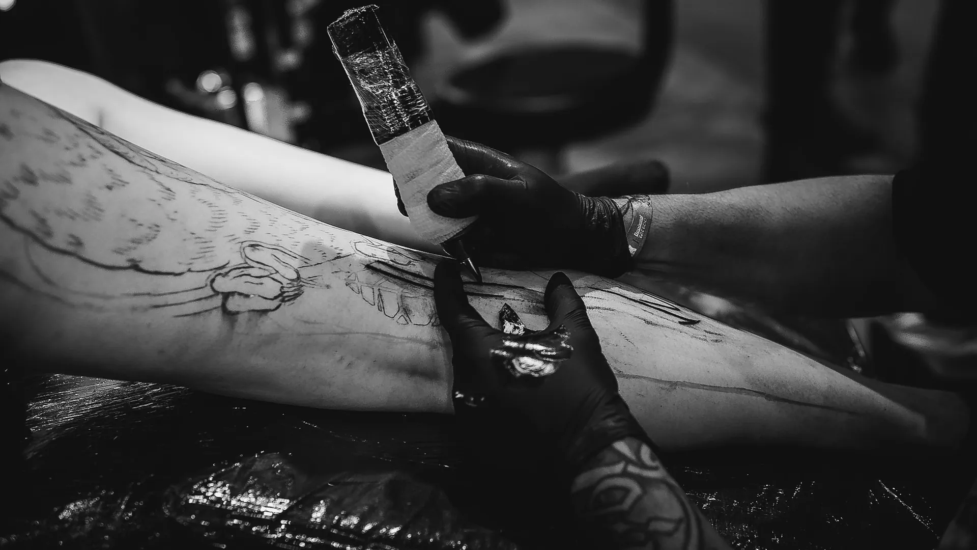 Mãos de luva preta conduzindo a máquina de tatuagem sobre o estêncil na coxa, em sessão no estande da The VOID
