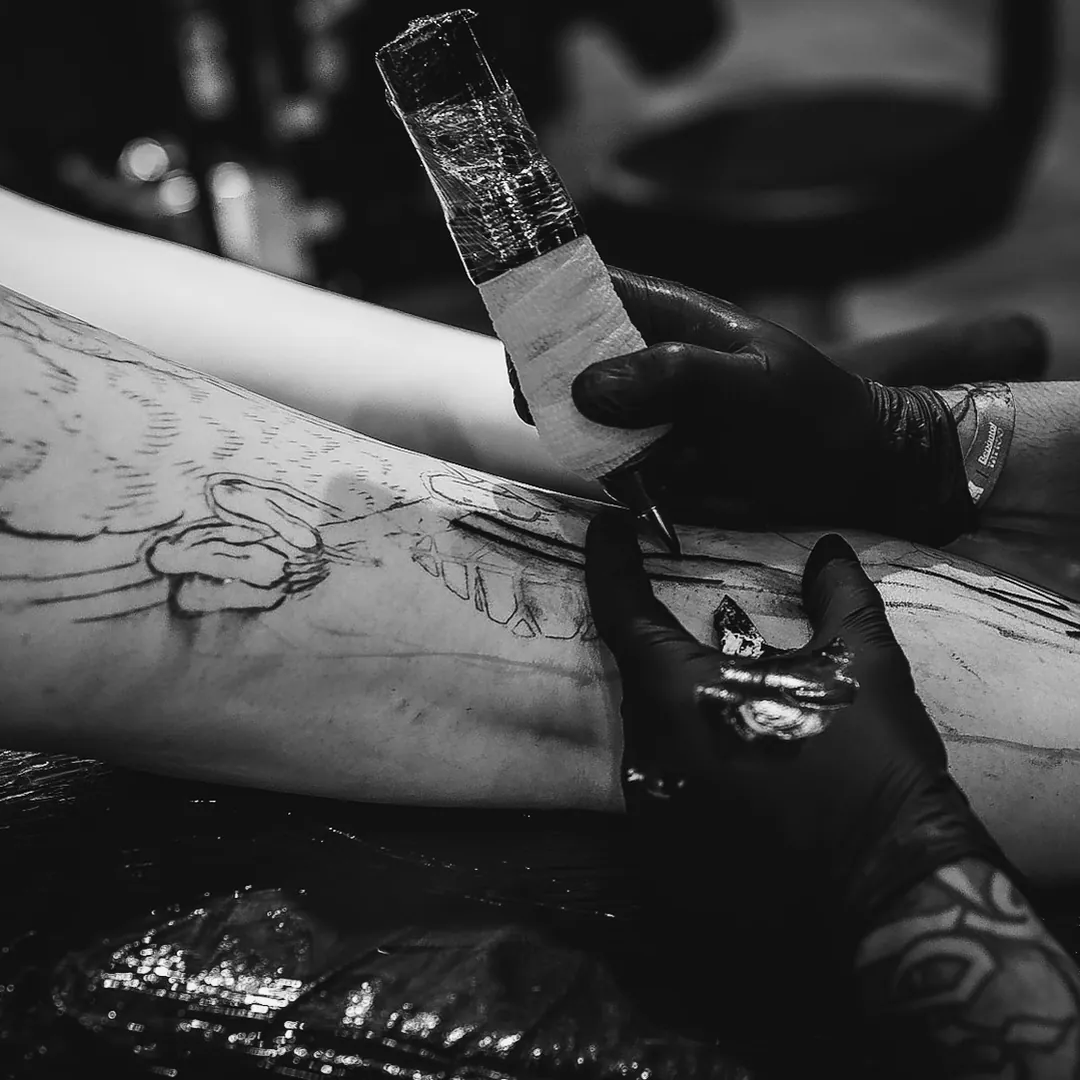 Mãos de luva preta conduzindo a máquina de tatuagem sobre o estêncil na coxa, em sessão no estande da The VOID