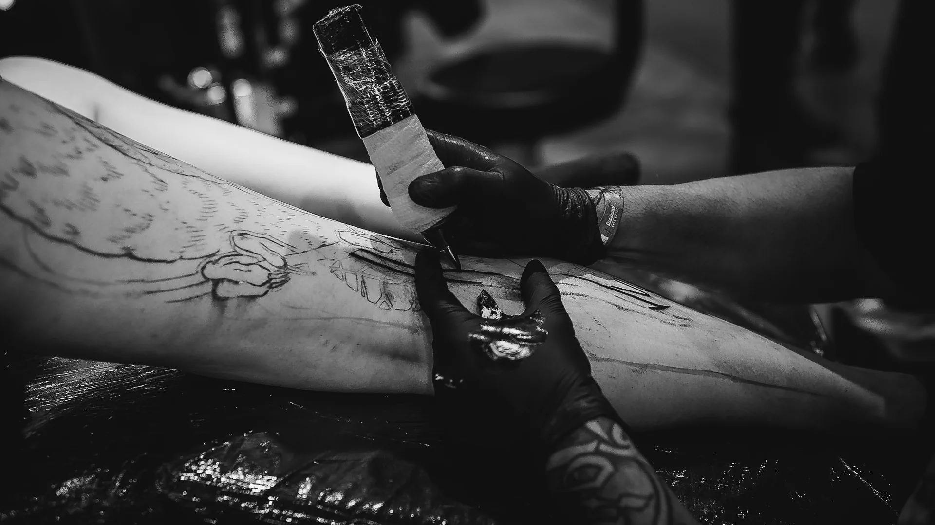 Mãos de luva preta conduzindo a máquina de tatuagem sobre o estêncil na coxa, em sessão no estande da The VOID
