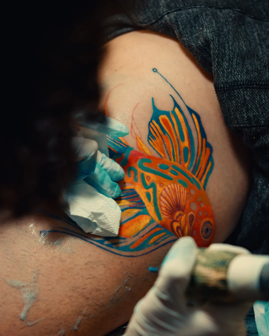 Máquina de tatuagem preenchendo um peixe colorido em laranja e azul na pele, mão com luva
