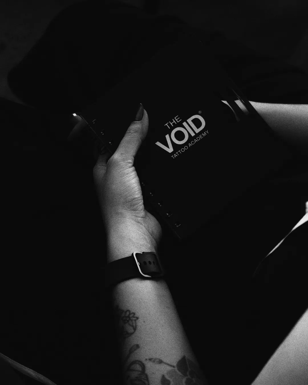 Mão tatuada segurando caderno preto com o logotipo da The VOID Tattoo Academy
