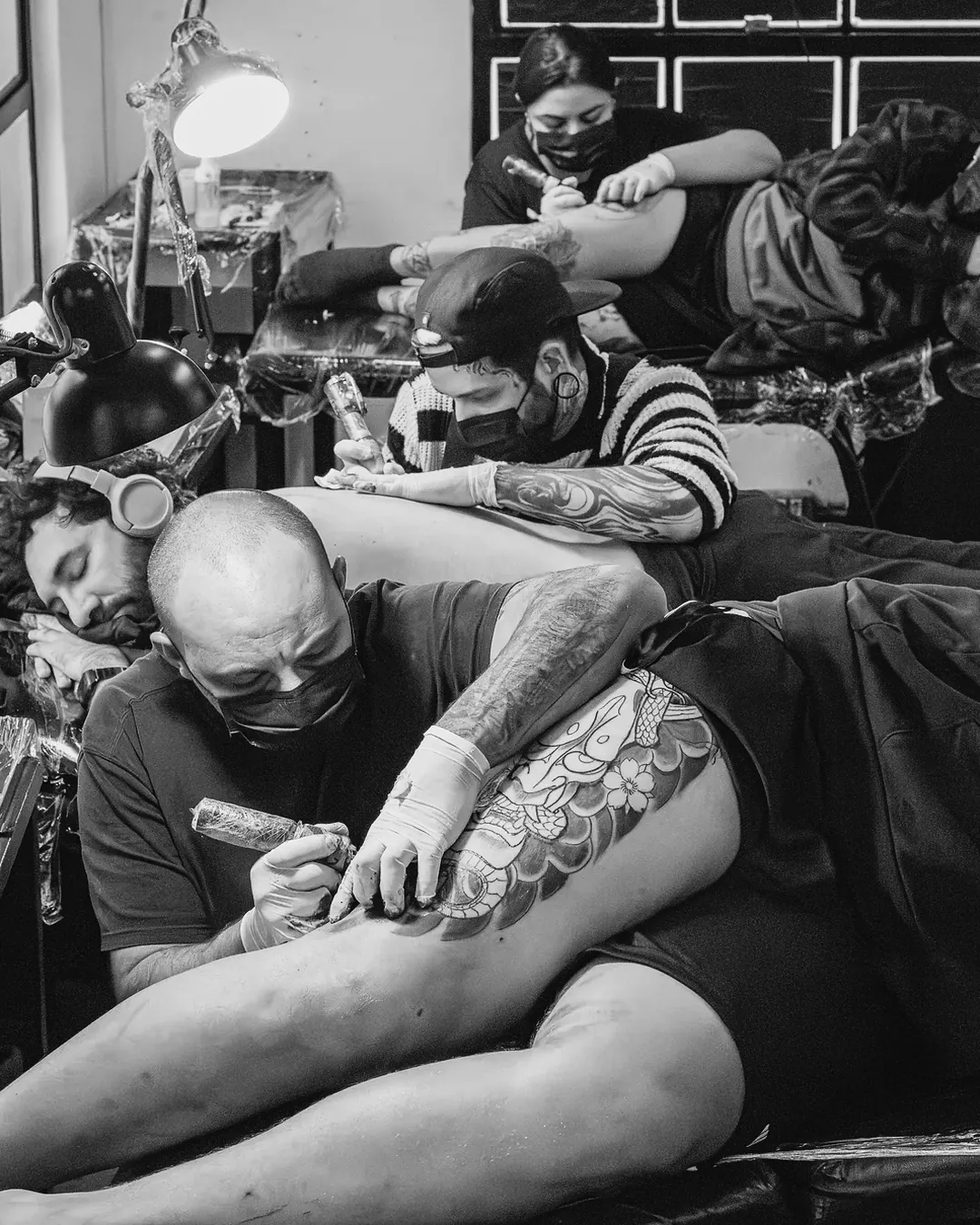 Professor e aluna de máscara tatuando juntos um cliente deitado de lado na escola