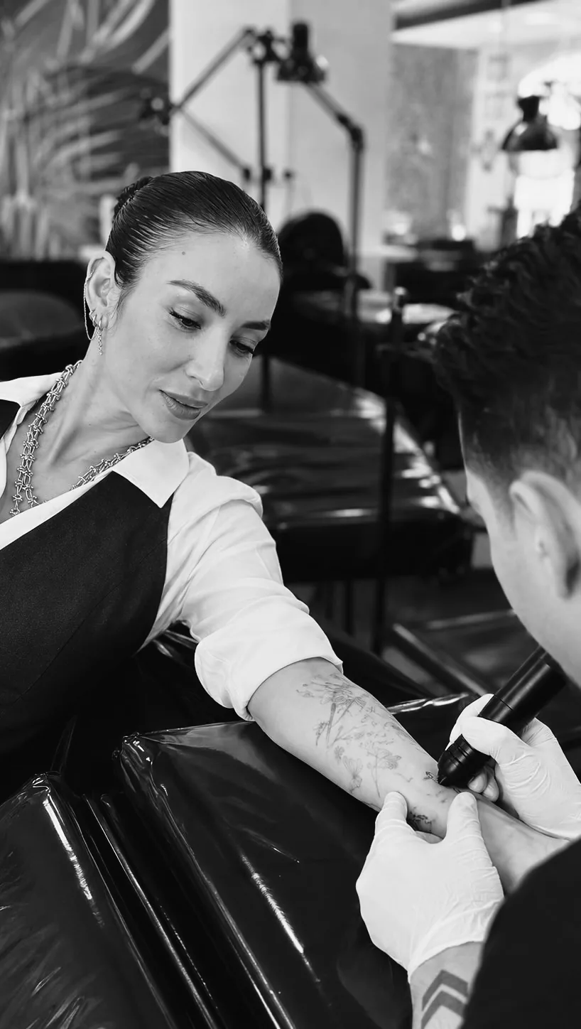 Professora orientando de perto um aluno que tatua o antebraço de outra pessoa