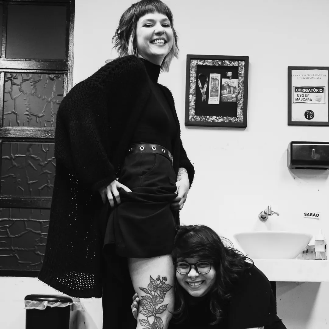 Aluna e tatuadora sorrindo com tatuagem cicatrizada na coxa, adesivo VOID na parede