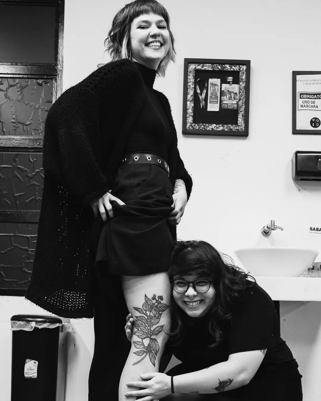 Aluna e tatuadora sorrindo com tatuagem cicatrizada na coxa, adesivo VOID na parede