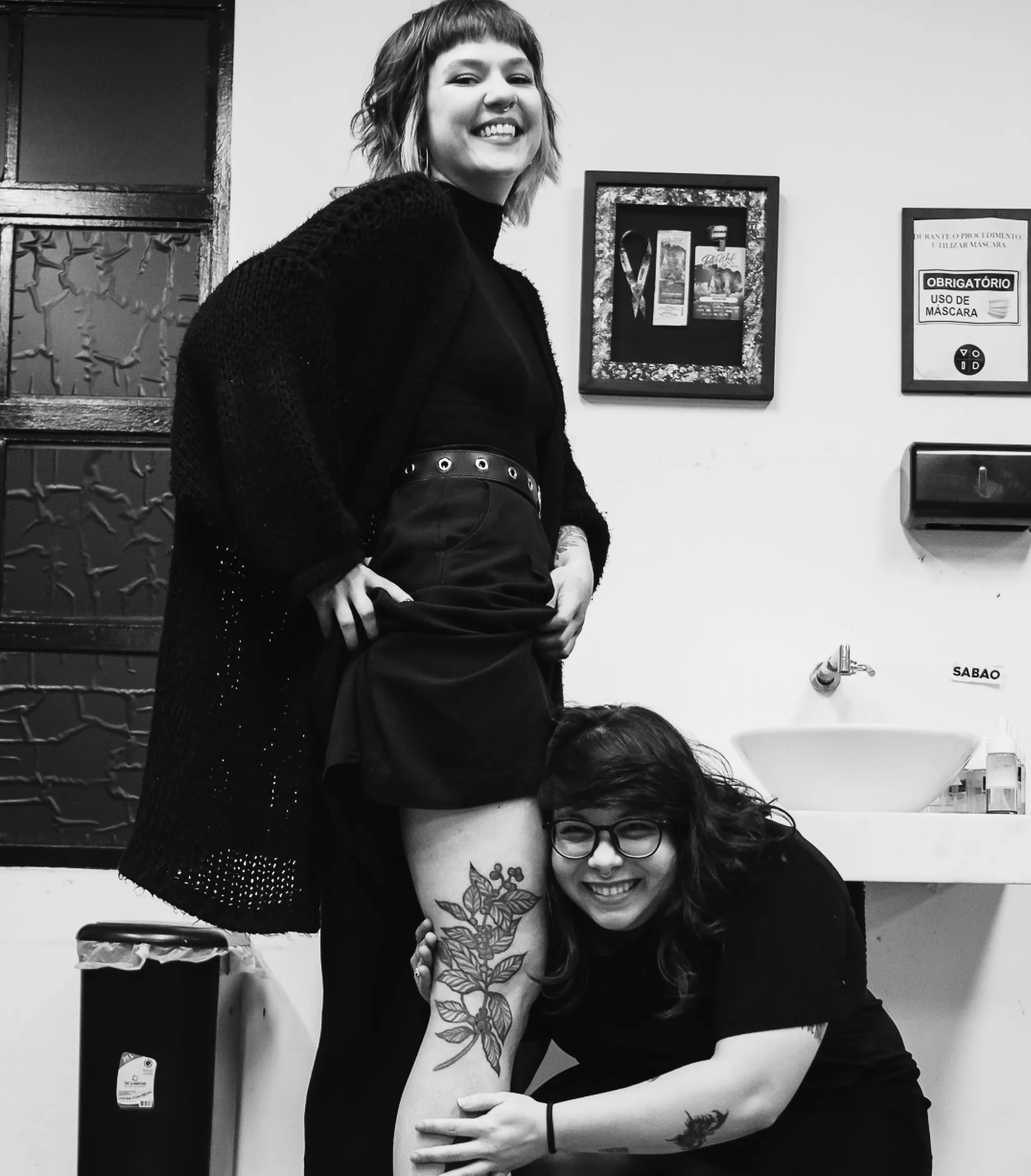 Aluna e tatuadora sorrindo com tatuagem cicatrizada na coxa, adesivo VOID na parede