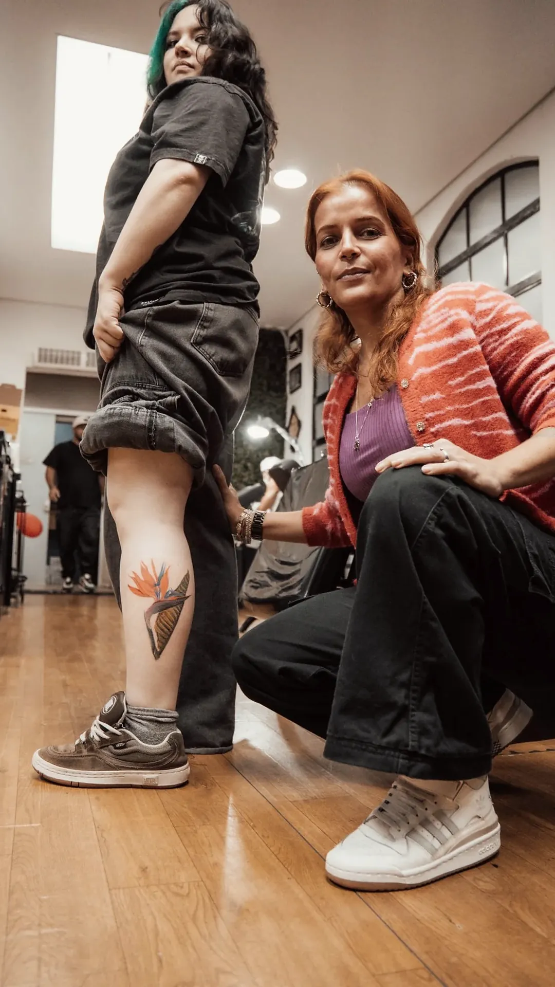 Aluna Isis agachada ao lado da cliente com a ave-do-paraíso tatuada, corredor da escola